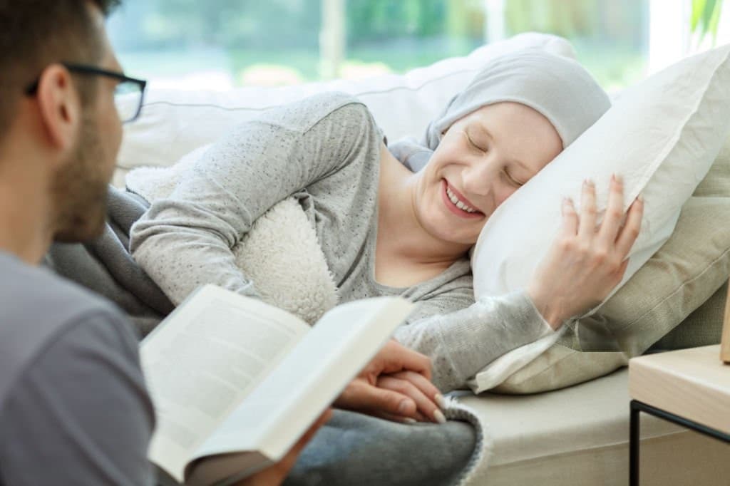A patient in a headscarf resting on a sofa while a visitor reads aloud