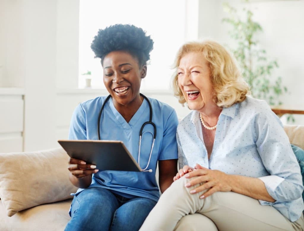 A nurse and an older patient laughing together over a tablet at home